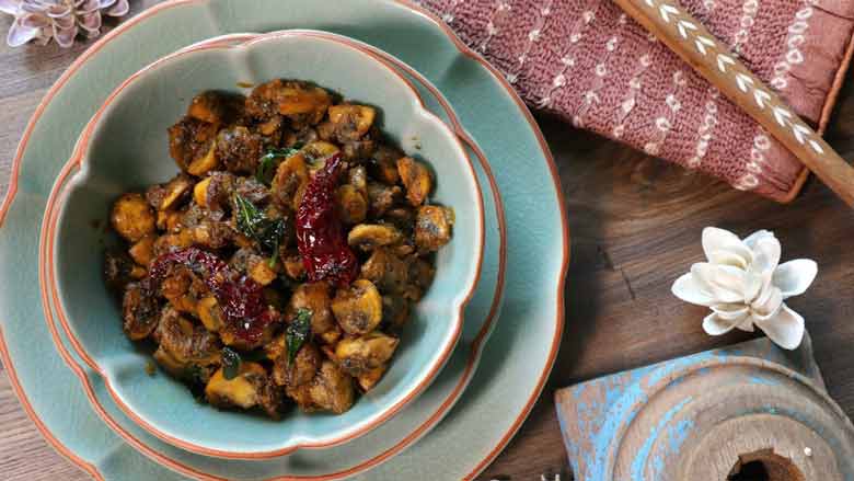 A top shot of mushroom pepper fry in a teal ceramic bowl with a terracotta coloured rim on two plates of the same colour. A copper colour tie-dye fabric, shell flowers, inlaid wooden spoon, and a blue wooden piece lie in the background - Mushroom Pepper fry Recipe
