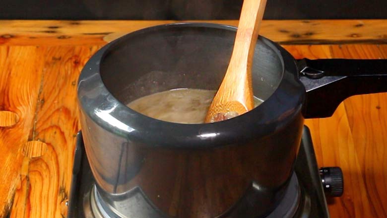 A spatula stirs mutton, spices, and water in a pressure cooker before sealing the lid. The black pressure cooker is on an electric stove on a pine wood countertop - Yakhni Recipe