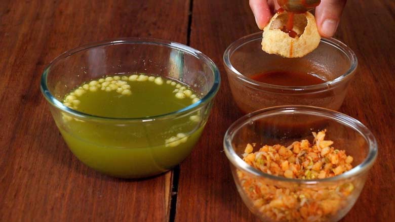 A hand is holding a puri while meetha chutney is being spooned into the puri. Glass bowls of potato mixture, meetha chutney, and teekha pani are seen on a wooden table - Pani Puri Golgappa Recipe