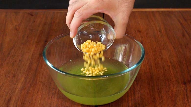 A hand is seen tipping boondi from a small glass bowl in to a large glass bowl half full of teekha pani on a wooden surface - Pani Puri Golgappa Recipe