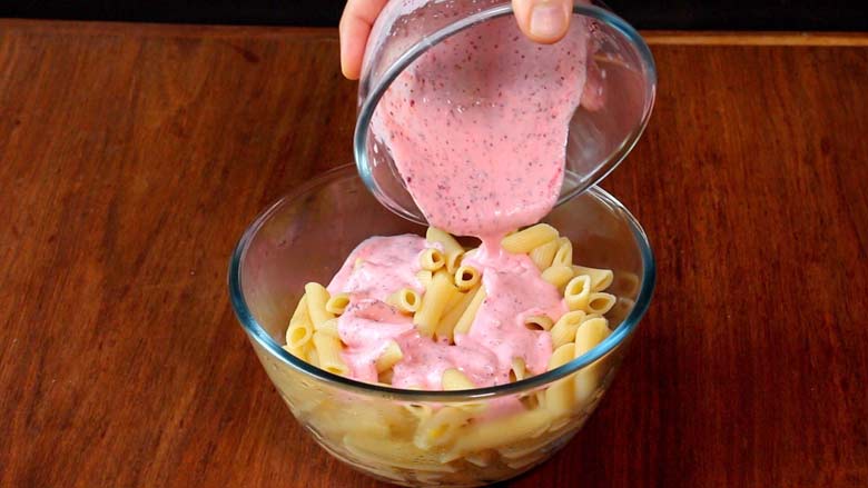 Boiled penne pasta is placed in a large glass bowl while a yogurt sumac mixture is being poured over it. The glass bowl lies on a natural wood table - Pasta with Herb Butter Sauce Recipe