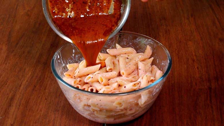 Prepared Browned Butter Sauce with sumac is being poured from a glass bowl onto penne pasta already coated with a yogurt sauce in a large glass bowl on a natural wood countertop - Pasta with Herb Butter Sauce Recipe