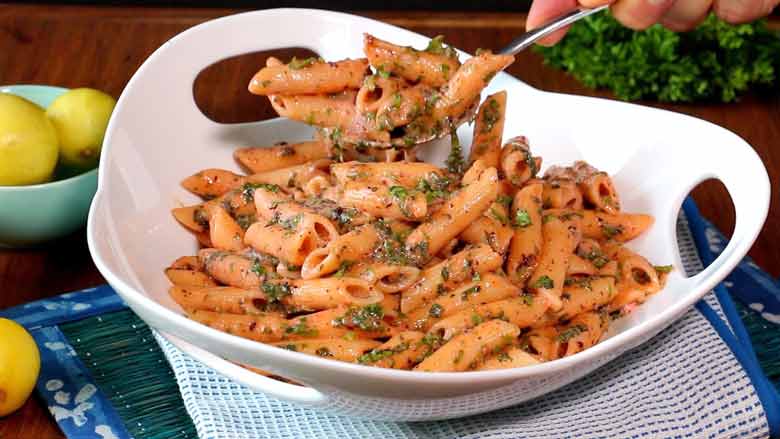 A spoon is seen picking up pasta from a large white ceramic bowl with three holes as handles on a white and blue mat on another emerald coloured mat with limes and parsley in the background - Pasta with Herb Butter Sauce Recipe