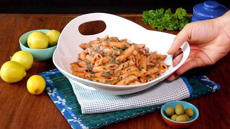 A hand is seen placing a large white ceramic bowl with three holes as handles with pasta with herb butter sauce on a white and blue mat on another emerald coloured mat with olives in a bowl in the foreground and limes and parsley in the background - Pasta with Herb Butter Sauce Recipe