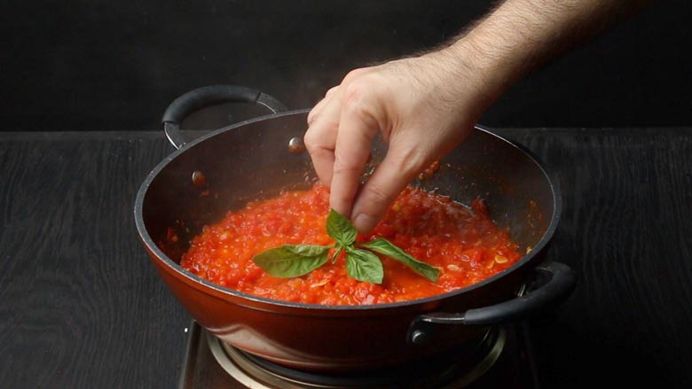 A hand is placing a sprig of basil on top of the tomatoes cooking on the pan. The pan is on an electric stove which is placed on a black counter top - Pizza Sauce Pasta Recipe