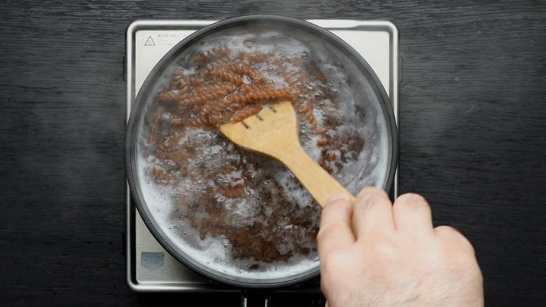 A hand is seen using a wooden spatula to stir the fusilli pasta in boiling water in a large black saucepan on an electric stove, on a black wooden counter top - Pizza Sauce Pasta Recipe
