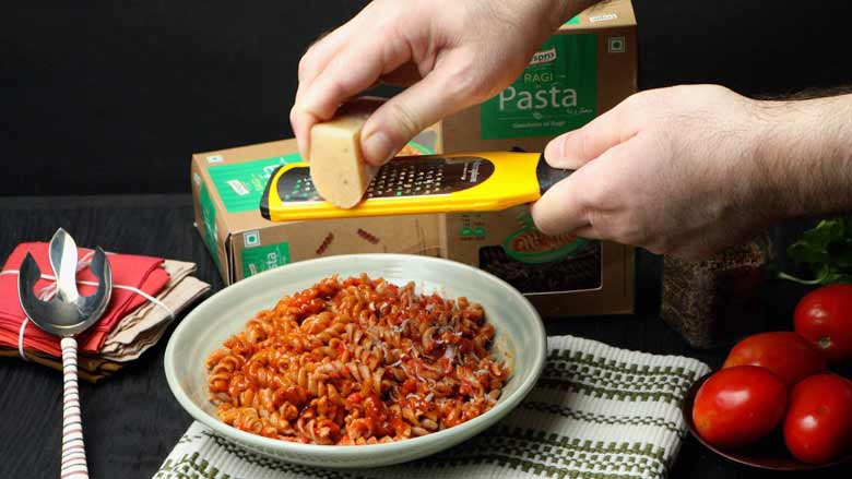 Two hands are seen grating parmesan cheese with a yellow grater over a bowl full of pizza sauce pasta with tomatoes in the foreground and a box of Fespro Ragi Fusilli Pasta in the background - Pizza Sauce Pasta Recipe