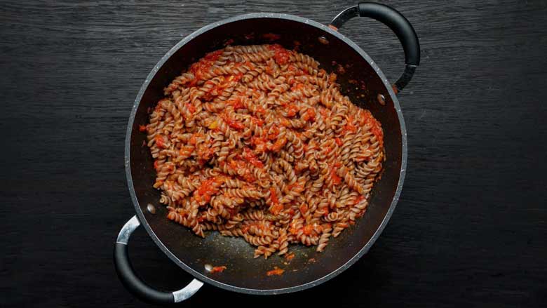 A top view of a non stick pan filled with ragi fusilli pasta in a marinara sauce on a black wooden counter top - Pizza Sauce Pasta Recipe