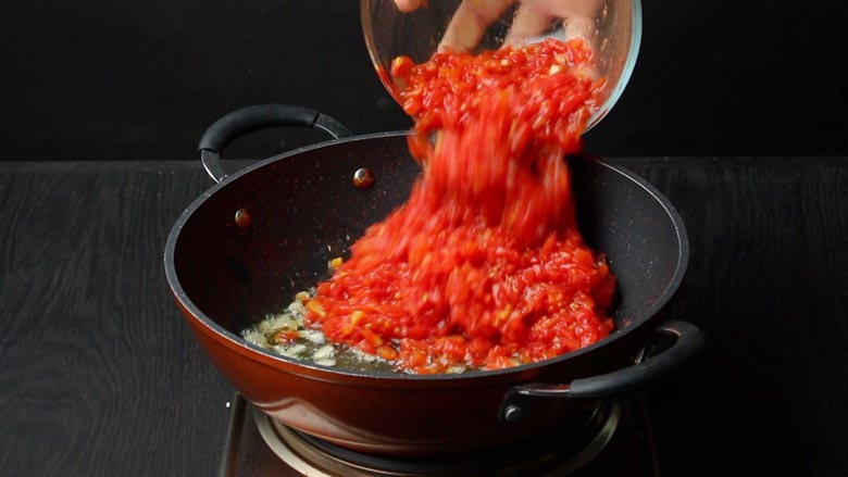 Blanched, peeled, and chopped tomatoes being added from a glass bowl to an orange and black non stick pan with garlic already sautéing in the pan - Pizza Sauce Pasta Recipe
