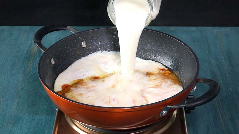 Milk is being poured from a glass jug into the semiya preparation in an orange and black non stick pan on a metallic stove on a blue wooden counter top - Semiya Payasam Recipe