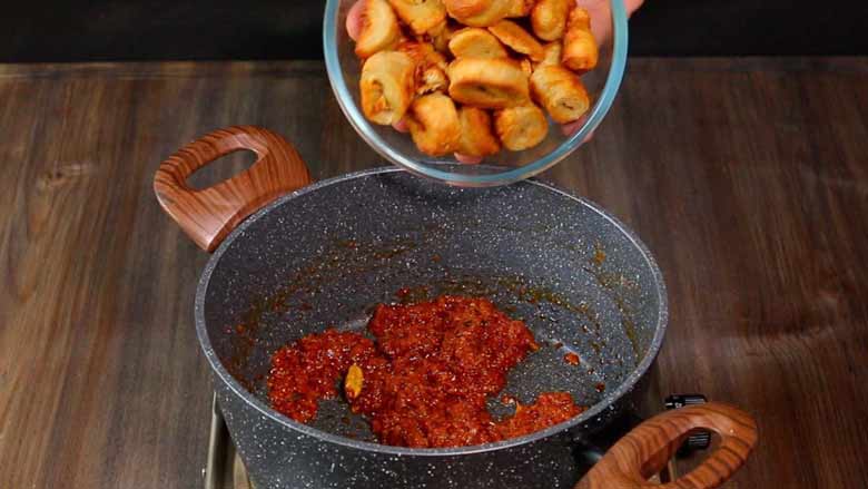 A glass bowl fulling of golden brown soya chaap is seen being tipped into prepared vibrant coloured rogan josh masala in a grey and white speckled non stick pan on a stove on a wooden counter - Soya Curry Recipe