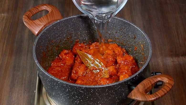 Water from a glass jug is being poured into the cooked soya chaap masala to thin out the curry in a non stick pan with 2 large wooden handles on a steel electric stove. On a wooden counter - Soya Chaap Curry Recipe