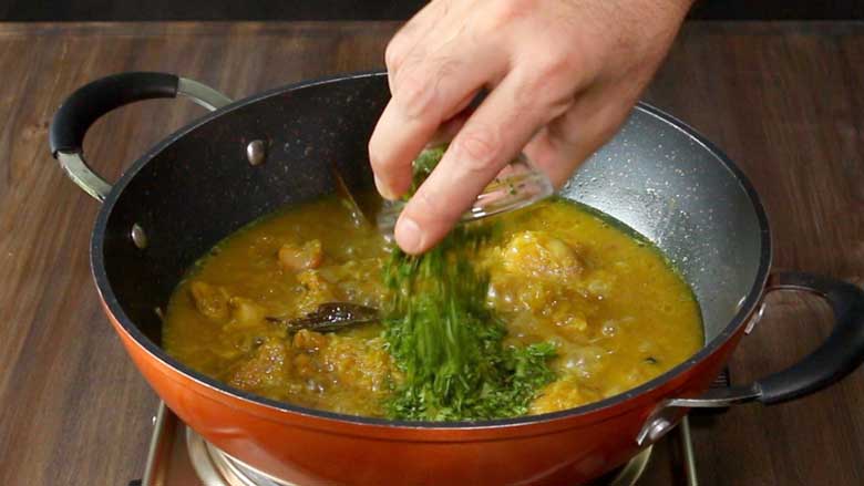 A hand is seen tipping fresh coriander leaves into the mirch kaliya being cooked in an orange and black pan with two handles - Spicy Meat Curry Recipe Mirch Kaliya