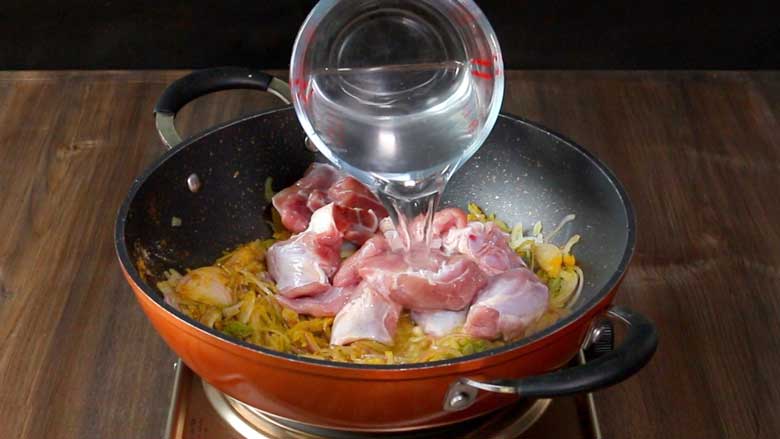 Water is being poured from a glass measuring cup over mutton and an onion mixture in an orange and black nonstick pan with two handles on a metallic electric stove on a wooden counter - Spicy Meat Curry Recipe Mirch Kaliya