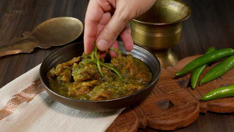 A hand is seen placing green chilli juliennes as a garnish over an easy mutton curry in a metallic brown ceramic bowl lying on an ecru and gold handwoven mat on a carved wooden platter with green chillies, a copper rice spatula, and a copper bowl in the background - Spicy Meat Curry Recipe Mirch Kaliya
