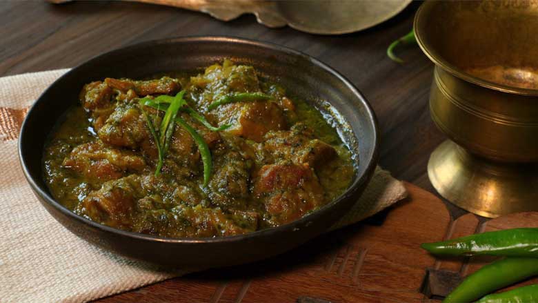 A side angled image of spicy mutton curry in a metallic brown ceramic bowl lying on an ecru and gold handwoven mat on a carved wooden platter with green chillies, a copper rice spatula, and a copper bowl in the background - Spicy Meat Curry Recipe Mirch Kaliya