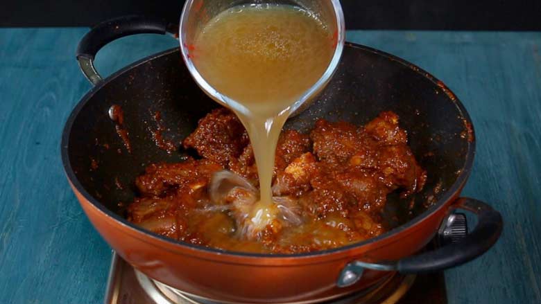 Meat stock being poured into a non-stick pan, with an orange coloured exterior with browned meat and other ground spices, on a metallic electric stove on a blue painted wooden table while making Rampuri Mutton Curry - Taar Gosht Recipe