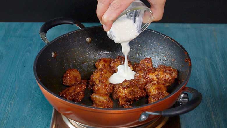 A hand is seen tipping a small glass bowl of yogurt into browned meat in a non-stick pan with an orange coloured exterior on a metallic electric stove on a blue painted wooden table with a black background while making Rampuri Mutton Curry - Taar Gosht Recipe