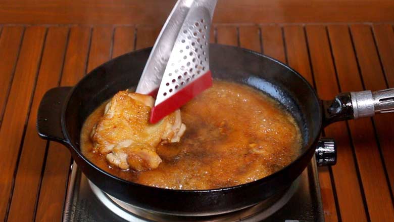 A stainless steel spatula with red silicon tips is seen placing a crispy fried chicken thigh back in the thickened teriyaki sauce in a black cast iron pan on a stainless steel electric stove on a slotted wooden surface - Teriyaki Chicken Recipe