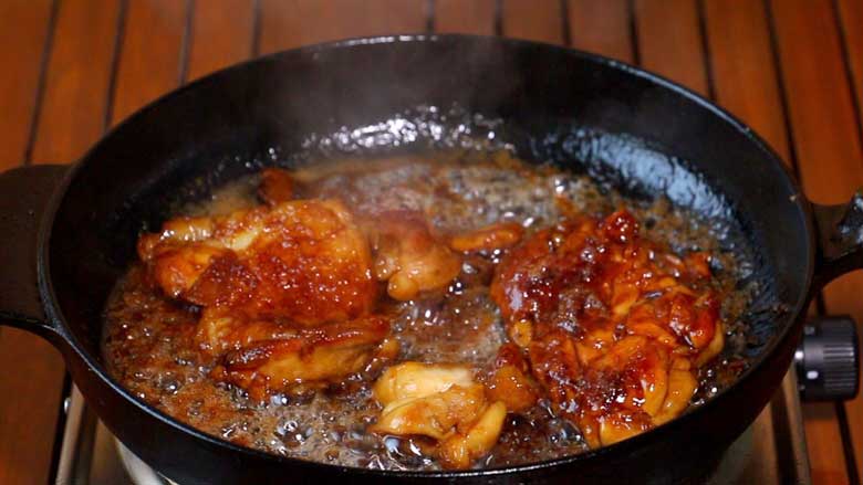 Boneless chicken thighs take on a beautiful golden gloss of the teriyaki sauce and begin to caramelise in a black cast iron pan. A slotted wooden surface can be seen under the pan - Japanese Teriyaki Chicken recipe
