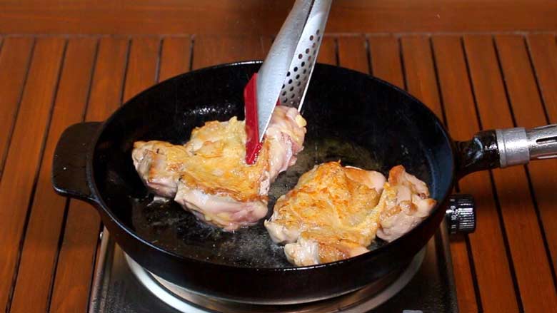 A spatula is seen turning a chicken thigh frying in a black cast iron pan. The two chicken thighs looks crisp and golden. The pan is on a metallic electric stove on a slotted wooden table - Teriyaki Chicken Recipe