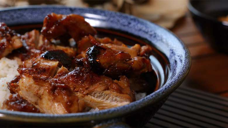 A close up shot of sliced Teriyaki Chicken on rice in a patterned blue and brown ceramic bowl. The chicken has a deep gloss and lustre of the teriyaki sauce. The bowl is lying on a slotted wood mat with chopsticks - Teriyaki Chicken Recipe