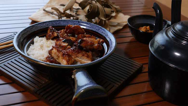 A side shot of Teriyaki Chicken slices placed on rice in a single handled patterned blue and brown ceramic bowl. The bowl is lying on a slotted wood mat with chopsticks visible and dried leaves in the background and a small black bowl of teriyaki sauce and a black antique kettle on the right on a slotted wooden surface - Teriyaki Chicken Recipe