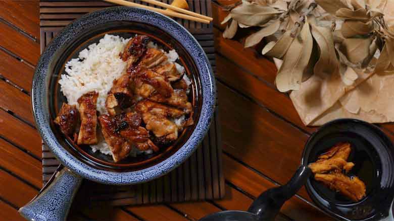A top shot of Teriyaki Chicken slices placed on rice in a single handled patterned blue and brown ceramic bowl. The bowl is lying on a slotted wood mat with chopsticks visible and dried leaves and a small black bowl of teriyaki sauce on the right on a slotted wooden surface - Teriyaki Chicken Recipe