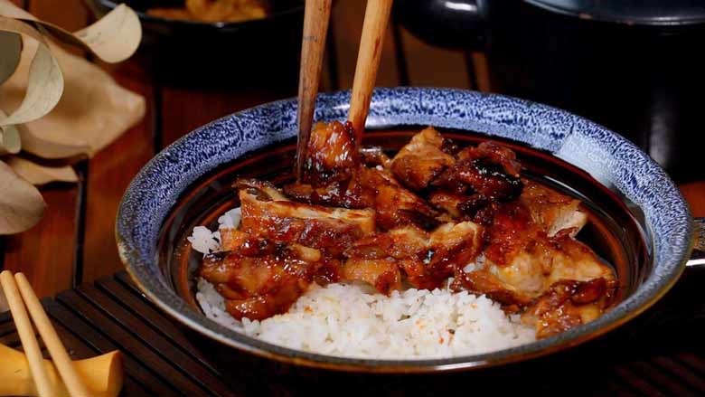 A close up shot of wooden chopsticks placing a sliced piece of Teriyaki Chicken in a patterned blue and brown ceramic bowl half filled with rice. The bowl is lying on a slotted wood mat with chopsticks in the foreground and dried leaves and a small black bowl of teriyaki sauce on a slotted wooden surface in the background - Teriyaki Chicken Recipe