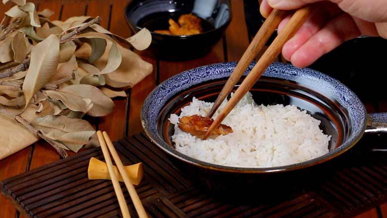 A hand is seen holding wooden chopsticks and placing a sliced piece of Teriyaki Chicken in a patterned blue and brown ceramic bowl half filled with rice. The bowl is lying on a slotted wood mat with chopsticks in the foreground and dried leaves and a small black bowl of teriyaki sauce on a slotted wooden surface in the background - Teriyaki Chicken Recipe