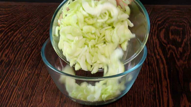 Cucumber that has been cut in half rounds or half moons is being transferred from a smaller glass bowl to a larger glass bowl lying on a heavily grained wood counter - Thai Cucumber Salad recipe