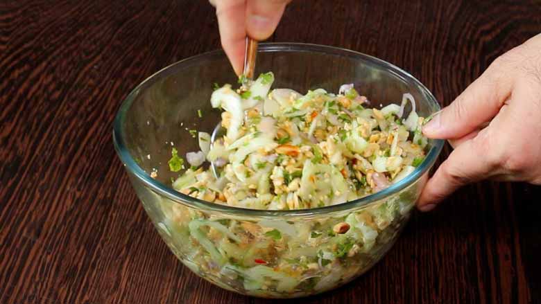 Hands are seen holding a glass bowl with sweet and sour cucumber salad ingredients and mixing the ingredients with a spoon. The glass salad bowl lies on a heavily grained wood table - Thai Cucumber Salad