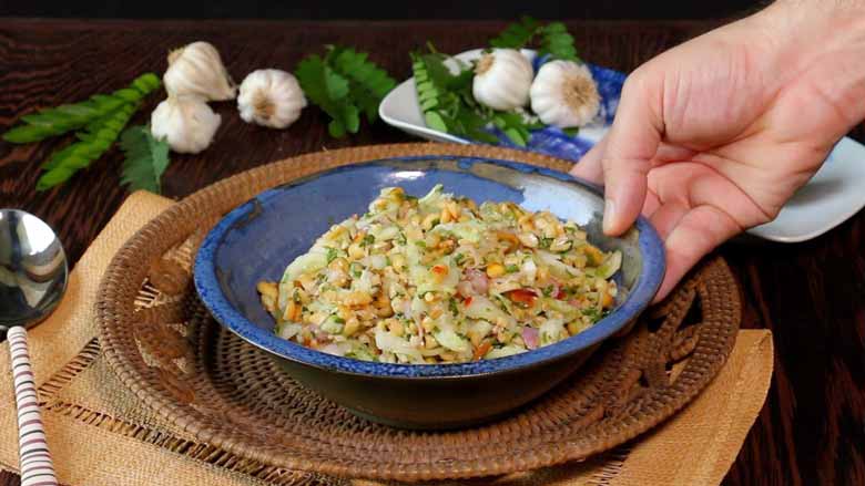 A hand is seen placing a blue and copper ceramic bowl filled with Thai cucumber salad on a woven cane platter lying on another cane mat with whole garlic with leaves and a tie dye ceramic platter in the background - Thai Cucumber Salad Recipe