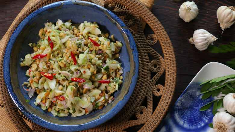 A clear top shot of the Thai cucumber salad with shallots and peanuts and topped with optional red chillies in a blue and copper ceramic bowl lying on a woven cane bowl with whole garlic heads, leaves, and a blue tie dye ceramic platter in the background- Thai Cucumber Salad Recipe