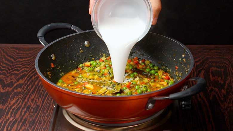 Thick coconut milk is being poured from a glass bowl into an orange and black non stick pan with cooked diced vegetables to make vermicelli pulao - Vermicelli Pulao Recipe