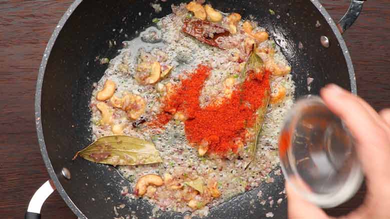 A hand is seen tipping red chilli powder from a small glass bowl into a large non stick kadhai with onions and other spices being sautéed in it - Vermicelli Pulao Recipe