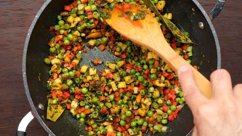 A colourful top shot of a hand holding a wooden spatula stirring diced assorted vegetables being sautéed in a black non stick kadhai - Vermicelli Pulao Recipe