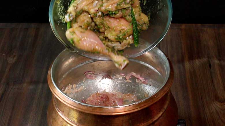 The marinated chicken is being tipped from a glass bowl into very lightly browned onions cooking in a copper handi - Village Style Chicken Biryani Recipe