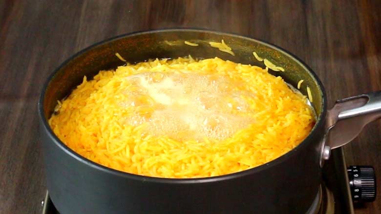 Rice that has been yellowed with turmeric is seen boiling and nearly ready in a grey nonstick pan with a single handle on a metallic electric stove on a wooden countertop - Village Style Chicken Biryani Recipe