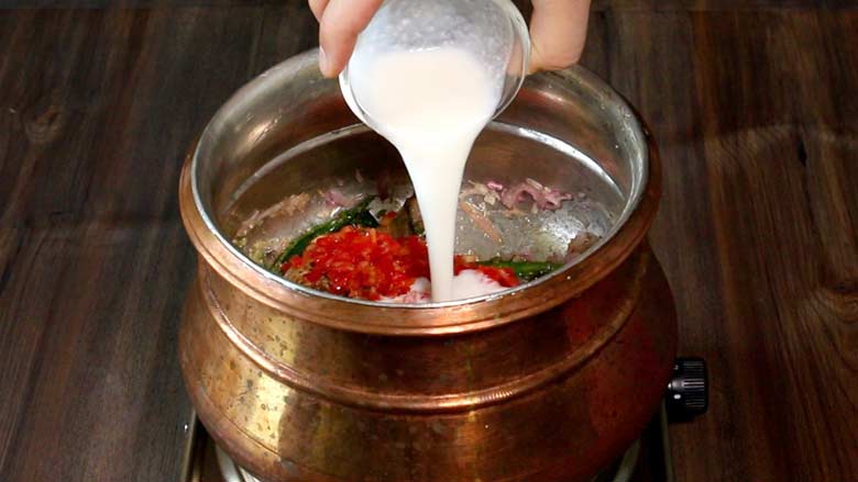 A hand is seen pouring yogurt from a small glass bowl into a copper handi where tomatoes have just been added. The copper handi is on an electric stove that lies on a wooden countertop - Village Style Chicken Biryani