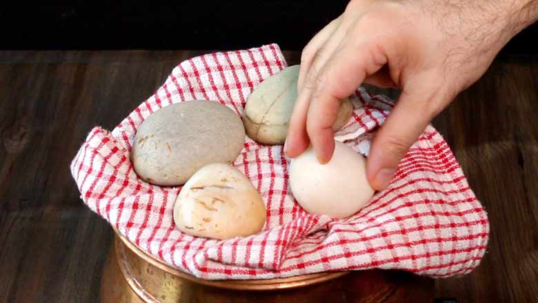 A hand is seen placing large riverbed stones on a red and white chequered cloth covered lid of a copper handi to give the chicken biryani dum - Village Style Chicken Biryani Recipe